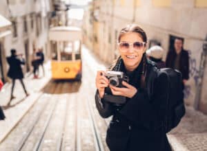 Smilende kvinde i storby. Hun har solbriller og står klar med sit kamera. I baggrunden en gul sporvogn.